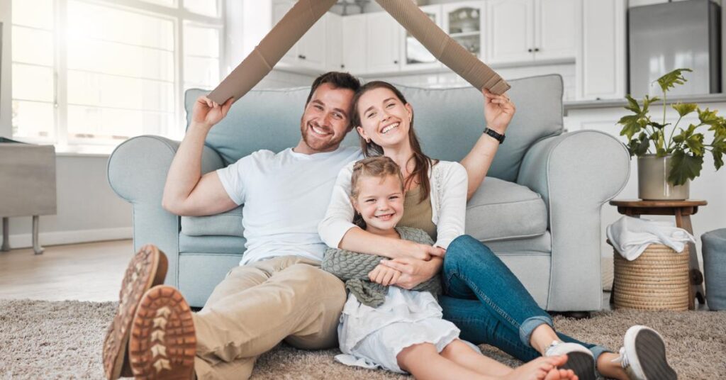 A smiling family of three sits on a rug in a bright living room, holding cardboard in the shape of a roof over their heads.