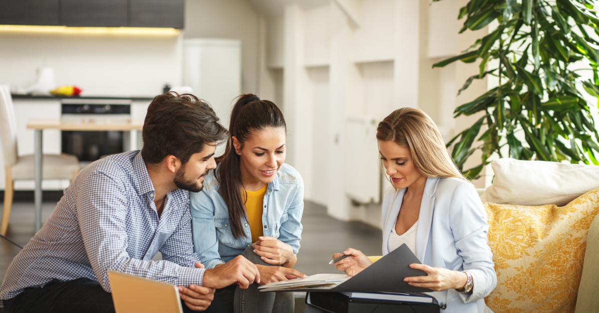 Three people sit in a living room, reviewing insurance documents together. A woman on the right is showing them a folder.