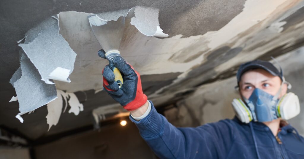 A person wearing a respirator is using a scraper tool to remove fire and smoke residue from a ceiling during a restoration.
