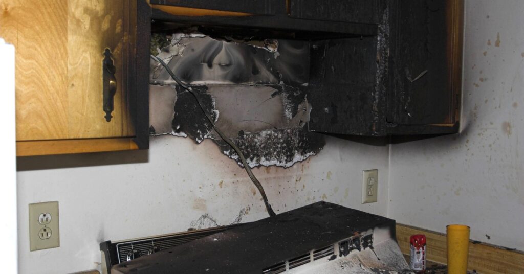 A stove and wooden kitchen cabinets blackened with fire damage and smoke residue after a kitchen fire.
