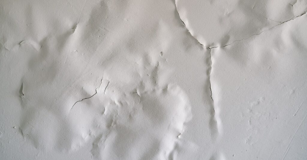 A close-up on a white wall with large cracks and bulges in the paint. The cracks run perfectly along the bulges.