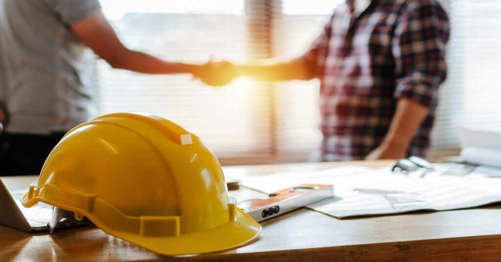 A yellow hard hat, a level tool, and some documents sit on a table as two people shake hands in the background.