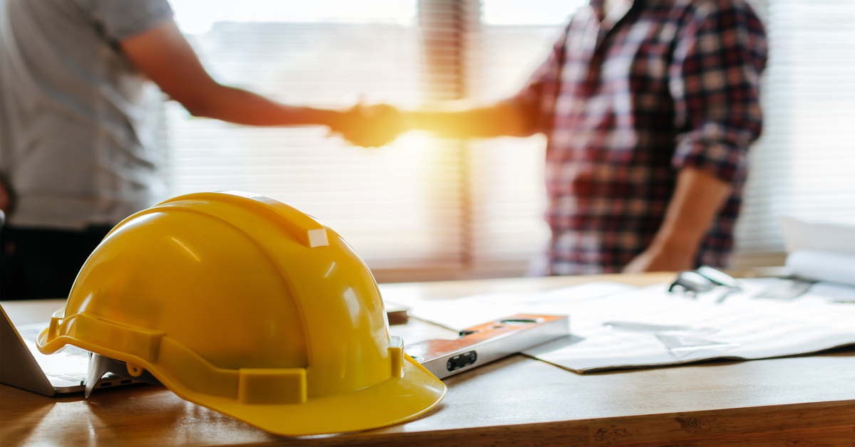A yellow hard hat, a level tool, and some documents sit on a table as two people shake hands in the background.