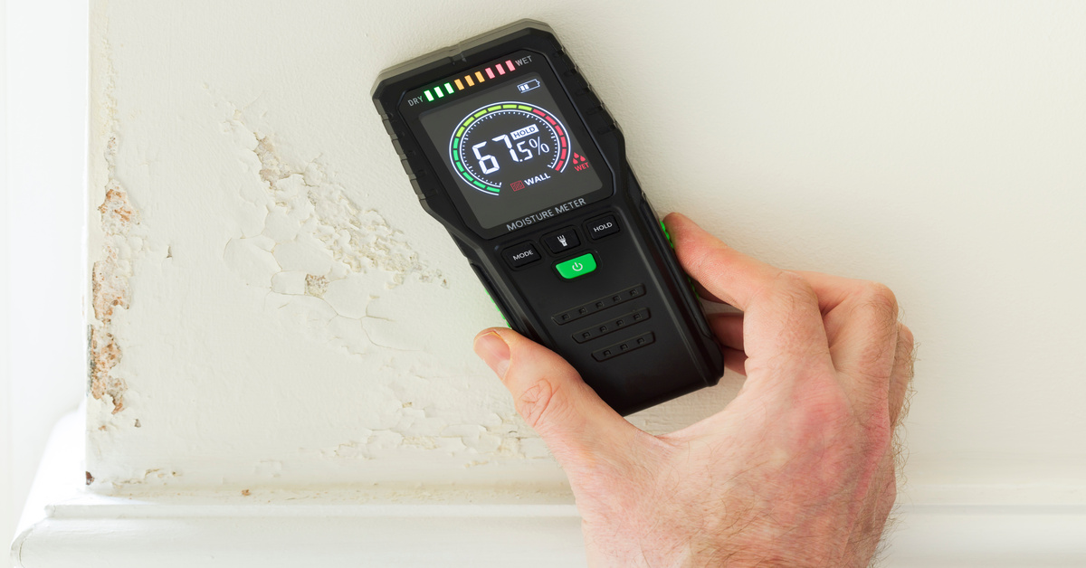 A person holds a moisture meter up to a wall to detect hidden water damage during a property restoration. It reads "67.5%".